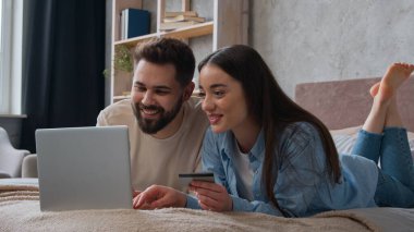Kafkasyalı çift yatakta yan yana yatıyorlar kredi kartı kullanıyorlar dizüstü bilgisayar bankacılığı uygulamasına şifreyi giriyorlar gülümseyen kadın ve erkek alışveriş siparişleri ev yatak odasında ödeniyor.