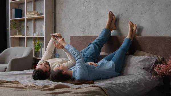 Carefree Caucasian couple in love lying down kick up feet on bed family weekend relaxing in bedroom man husband and woman wife use mobile phone application make internet order online shopping at home