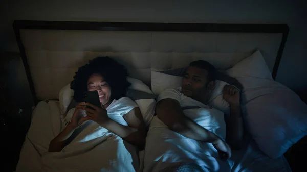 African American couple family in bed bedroom night angry frustrated man husband trying sleep offended on wife woman laughing loud using gadget addict disturb sleeping mobile phone addiction problem