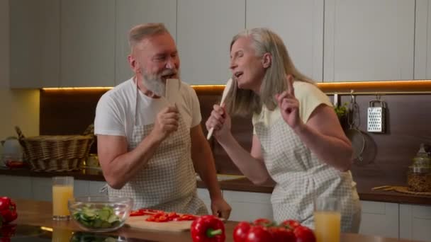 成熟した灰色の妻と夫の古いカップルの祖父母は 家庭のキッチンで歌う楽しい料理ランチ野菜サラダ引退コーカサス人男性の女性ダンスキッチンウェアスプーンの栄養で歌う音楽にダンス — ストック動画