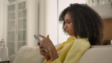 Sıkılmış Afrikalı Amerikalı etnik kadın cihaz bağımlısı kız internette sohbet ediyor aşırı sosyal medya bağımlılığı tembel, üzgün kadın ev sahibi ev telefonundan cep telefonuyla haberlere kayıyor.