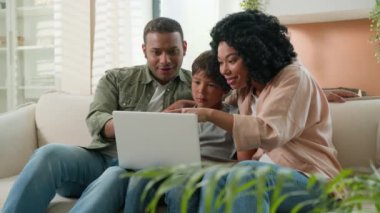 Mutlu Afro-Amerikan aileler anne baba ve beyaz çocuk çocuk çocuk çocuk çocuk dizüstü bilgisayar oyunu oynuyorlar eğlenceli çizgi filmlerin kazanmasını izliyorlar evdeki koltukta online zafer kutlaması yapıyorlar.