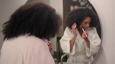 Güzel Afro-Amerikalı Etnik kadın banyo evinde aynadaki yansımaya bakıyor. Yüze pudra sürüp makyaj malzemesi hazırlıyor. Kozmetikle çıkmaya hazırlanıyor.