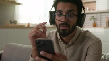 Mutlu gülümseyen Hintli adam, mutlu Arap adam kulaklıkla müzik dinliyor ev mutfağında dinleniyor cep telefonu müzik listesindeki sesi dinliyor kablosuz kulaklıklarda.