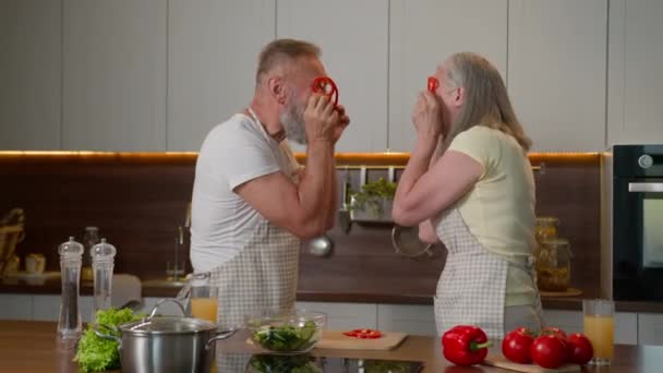 上了年纪的夫妇在厨房做沙拉 新鲜健康的蔬菜 有趣的是 成熟的女人和退休的白种人在厨房里拿着红辣椒 眼睛上挂着滑稽的眼镜 鬼鬼鬼祟祟的 — 图库视频影像