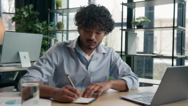 アラビアのラテン系男男性ビジネスマン インド人労働者 雇用主 執筆マネージャー オフィスデスクで書くビジネスノートを勉強 紙のノートブックの作業スケジュールでアイデアを書く ラップトップコンピュータを見る — ストック動画