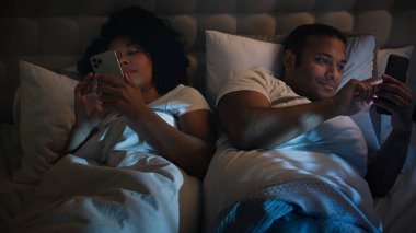 Gadgets bağımlısı internet bağımlısı Afro-Amerikan çift, aile erkeği, kız arkadaş, sevgili, gece evde rahat bir yatakta, cep telefonları kullanarak koca, birbirini görmezden gelen akıllı telefonlar.
