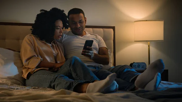 African American couple man and woman husband wife boyfriend girlfriend using mobile phone together lying in bed at night enjoying relaxing scrolling smartphone technology watching video social media