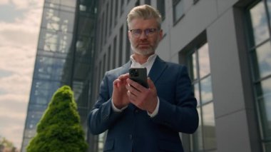 Gülümseyen orta yaşlı işadamı şehrin dışında ofis yakınında cep telefonu kullanıyor kameraya gülümse mutlu yetişkin iş adamı işvereni akıllı telefon simsarı emlakçısı