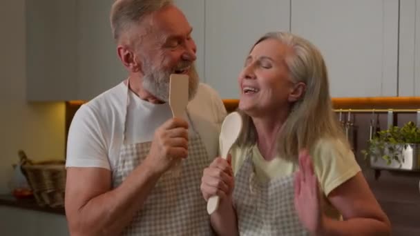 退休的中年白人家庭老两口在家里厨房唱卡拉Ok快乐有趣的夫妇灰发男人和女人在厨房里唱歌餐具汤匙跳舞有趣的烹调 — 图库视频影像