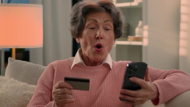 ハッピーシニア満足の年齢の白人女性は喜んでクレジット バンク カードが付いている自宅のソファーで引退した古い成熟した女性は成功した決済の携帯電話のオンライン バンキングのお金賞を喜びます — ストック動画