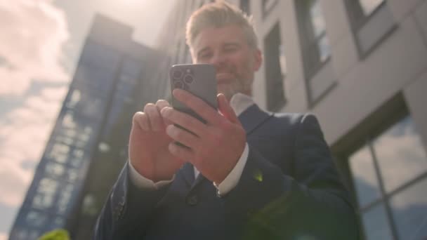 太陽の下で屋外で携帯電話を使用して年配のビジネスマンの笑顔 ハッピーシニア成人ビジネス男性ユーザー雇用主 携帯電話のテキスト スマートフォン オンラインチャット都市ビルディング Sunbeam — ストック動画