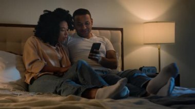 Afrika kökenli Amerikalı çift karı koca sevgili cep telefonunu birlikte kullanıyor geceleri yatakta uzanıyor akıllı telefon teknolojisini dinlendirip video sosyal medya izliyor.