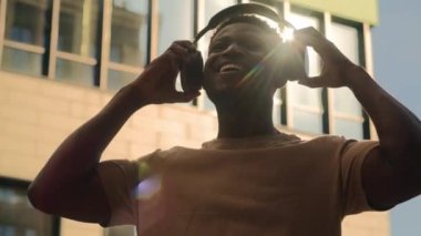 Mutlu gülümseyen Afrikalı Amerikalı, neşeli, gülümseyen adam, kablosuz kulaklık takıyor, müzik dinliyor, açık havada güneş ışığı yolcusu, şehirde eğleniyor, müzik dinletiyor.