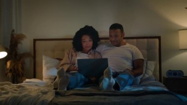 Afro-Amerikalı çift, aile babası ve kadın, gece yatağında, karanlık yatak odasında dizüstü bilgisayar izliyorlar, film izliyorlar, romantik randevu, aşk ilişkisi video alışverişi yapıyorlar.