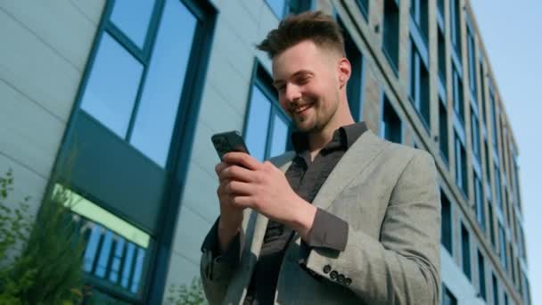 スマートフォン上のSmsメッセージを送るオフィスの建物の近くの携帯電話の歩道を使用してソーシャルネットワークをブラウジングするスマイリングペンシアンのビジネスマンビジネスマン起業家 — ストック動画