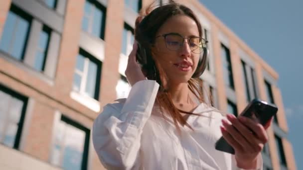 ヘッドフォンの幸せな女性は オフィスビルの近くの日光で屋外で音声ミュージックを聞く ケアフリーの女の女性 モバイルアプリケーションを使用して 都市の通りで休むお気に入りの曲の踊りを楽しむ — ストック動画