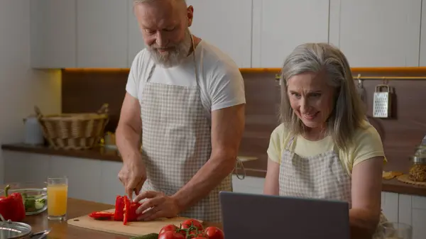 Caucasian retired couple in chefs aprons smiling cooking at home kitchen middle-aged woman wife using laptop culinary recipe old man husband cut pepper fresh vegetable salad family dinner healthy food