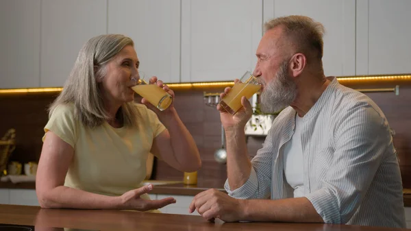Happy middle-aged old senior couple Caucasian family woman and man drinking orange juice at kitchen home cheerful talk spouses retired wife and husband drink fresh fruit healthy nutrition healthcare