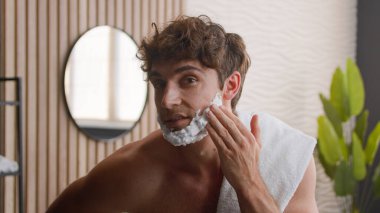 Close up Caucasian bearded male funny man guy looking at camera beauty routine morning hygiene procedure bathroom apply smear face white foam facial shaving gel cosmetic prepare for beard shave bath clipart