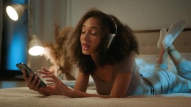 Afro-Amerikalı etnik kadın rahat bir yatakta uzanıyor. Kablosuz kulaklıkla müzik dinliyor. Yatak odasında dinleniyor.