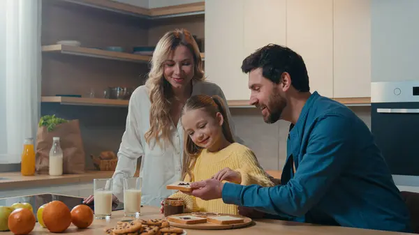 快乐的白人家庭一起准备早餐在家里交谈厨房里妈妈拿着眼镜和牛奶小女孩准备烤面包和巧克力面食和爸爸送来的食物 — 图库照片