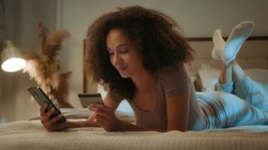 Afrikalı Amerikalı genç bayan müşteri kredi kartı kodunu girin cep telefonlu kadın alışverişçi internet alışverişi için ev yatak odasında dijital bankacılık kullanarak online alışveriş yapmak için
