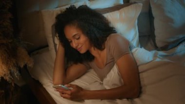 Gece bağımlısı Afrikalı Amerikalı genç bir kız, yatakta uzanmış akıllı telefon kullanıyor. Karanlık yatak odası, sosyal ağlar, mesajlar, uygulamalar, cep telefonuyla sohbet, yatmadan önce gülümseyen kız eğlencesi.