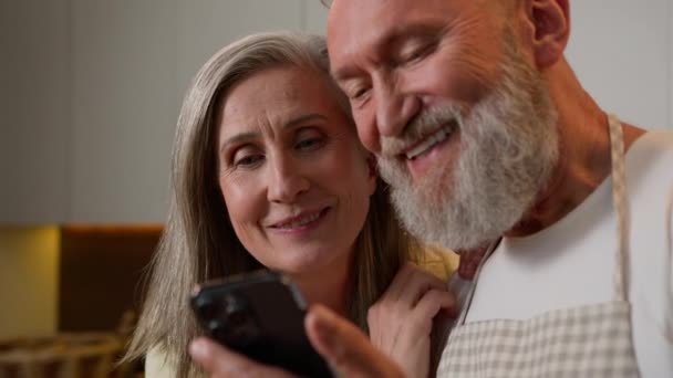 キッチンで笑う幸せな古い古いコーカサス家族を閉じる 面白いモバイルアプリを使用して成熟したカップルの祖父母 ニュースを読みます 引退した夫と女性の妻 オンラインでショッピング — ストック動画