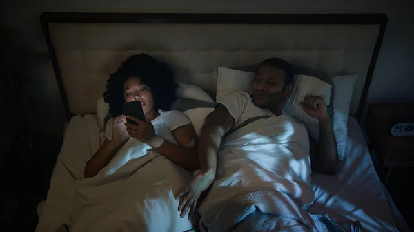 Irritated African American man boyfriend trying sleep husband angry push gadget addict wife internet addicted girlfriend woman scrolling mobile phone social media at night in bed couple family problem