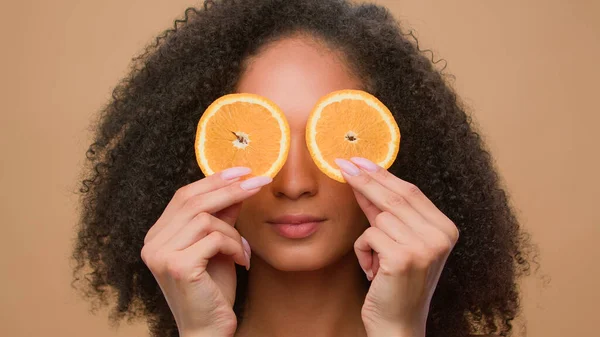 African American girl covering eyes with slices of citrus peeping blink eye woman hold two half of orange cosmetology dermatology skin care haircare dieting eco vitamin cosmetic ad beige background