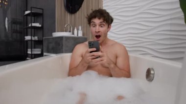 Banyoda sabunlu köpükle rahatlayan çıplak bir erkek banyo küvetinde dinleniyor. Şaşırmış görünüyor. Akıllı telefon uygulaması cep telefonunda iyi haberler okuyor.