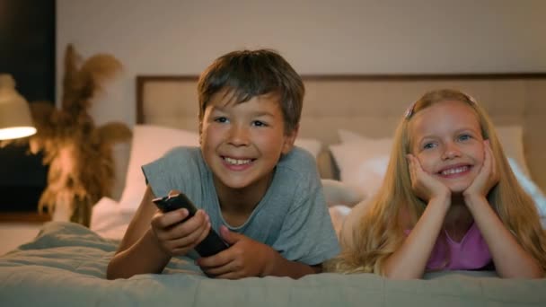 幸せな笑顔小さな慎重な子供の少年少女の兄弟姉妹は一緒に寝室のベッドに横たわってテレビを見ている子供の兄弟の腕時計漫画は家で笑って楽しんでいるリモート コントロールを保持します — ストック動画