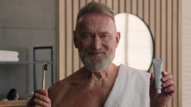 年长面带微笑的白种人中年男子退休男子白发苍苍的养恤金领取者在浴室拿着牙刷和牙膏牙齿保健日常卫生微笑 — 图库视频影像