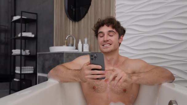 白人男性 裸の筋肉の若い男性 リラックスして浴室でリラックス 浴室バスタブ 会話ビデオ通話会議 オンラインチャット 携帯電話 ソーシャルメディアストリーミング — ストック動画
