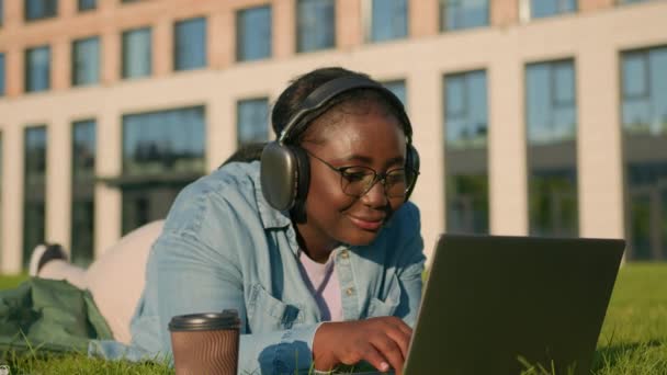快乐的女学生非洲裔美国女人躺在草坪上 躺在绿草上 耳机里听着音乐 耳机里有计算机听着歌曲 笔记本电脑在户外学习 — 图库视频影像