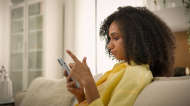 Sıkılmış Afrikalı Amerikalı etnik kadın cihaz bağımlısı kız internette sohbet ediyor aşırı sosyal medya bağımlılığı tembel, üzgün kadın ev sahibi ev telefonundan cep telefonuyla haberlere kayıyor.
