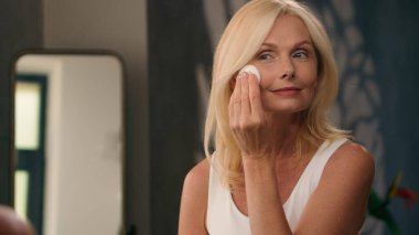 Beyaz, orta yaşlı, güzel bir kadın banyoda aynaya bakıyor. Yaşlanma önleyici toz sürüyor. Cilt kırışıklıkları var.