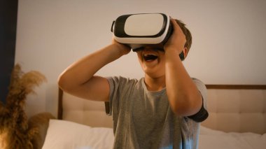 Komik hevesli çocuk okul çocuğu yenilikçi dijital VR gözlükler kullanarak video oyunu oynuyor. Meta-evren dünyasında 3D AR teknolojisi kullanıyor. Evde yatağın üzerinde sanal gerçeklik gözlüğü takıyor.