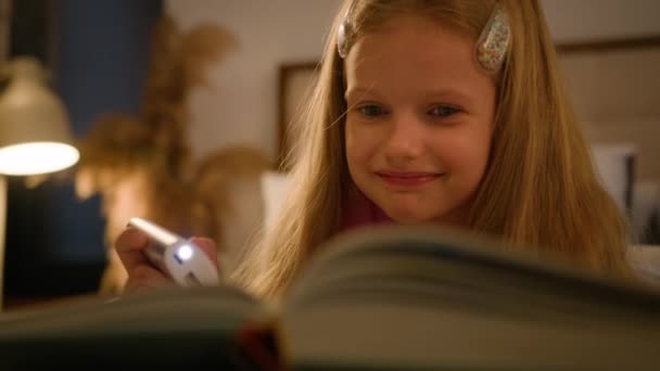 幸せな笑顔小さな慎重な子供の少女は夜に懐中電灯で本を読んで ベッドでリラックスして家でかわいい子供が楽しむおとぎ話を読みます ストーリーブックおかしな物語 — ストック動画