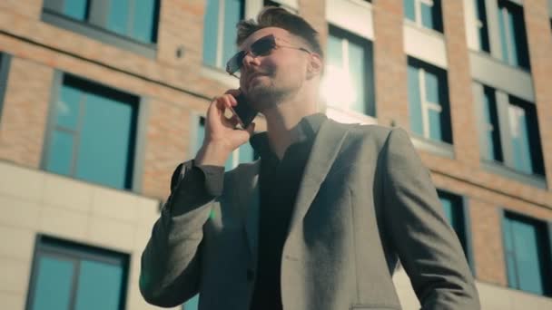 在阳光下的写字楼附近 戴着太阳镜的成功的高加索商人在户外对着手机讲话 男性创业者用智能手机答话进行远程交流 — 图库视频影像
