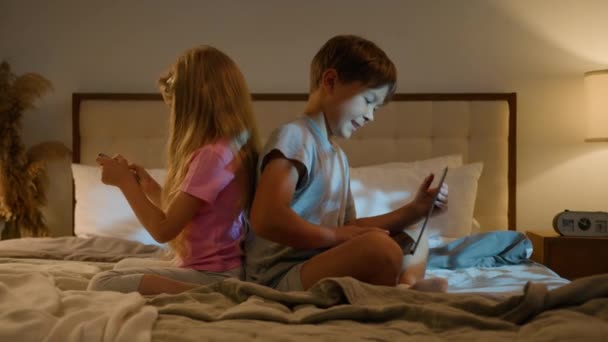 幸せな慎重な子供の少年少女の兄弟姉妹の友人はラップトップおよび電話の若い世代のインターネット技術の依存の概念を使用して自宅の夜にベッドで一緒にオンラインビデオゲームをする — ストック動画