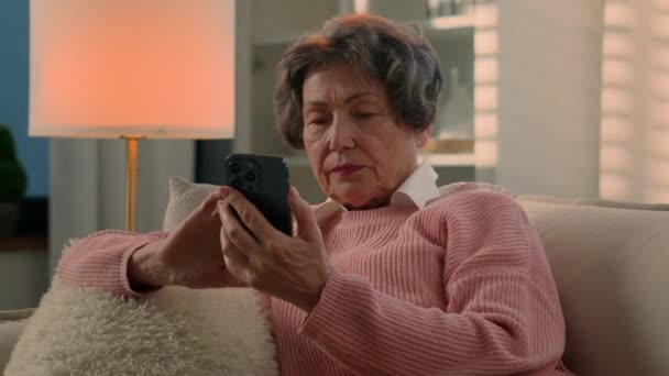 コーカサス古い女性祖母シニア女性 成熟した退職した女性 現代ハイテクスマートフォンを真剣に夜の家で読書ニュース ソファーショッピングアプリで携帯電話のインターネットサービスをスクロール — ストック動画