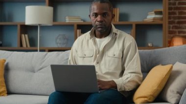 Afro-Amerikalı işadamı işadamı. Ev koltuğunda dizüstü bilgisayarda yazan müşterisi uzaktan kumandayla internet bilet alıyor. Oda kanepede poz veren gülümseyen etnik iş adamı.