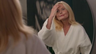Kafkasyalı yaşlı, olgun, bornozlu güzel kadın banyoya geliyor aynadaki yansımaya bakıyor doğal makyaj şampuanı saç bakımı sabah güzellik duştan sonra yaşlanma karşıtı rutin cilt bakımı