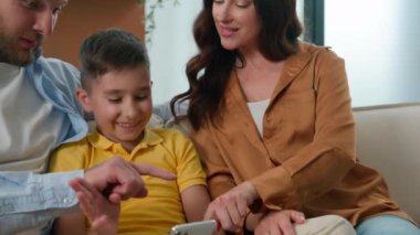Kafkasyalı mutlu bir aile babası, annesi ve çocuğu koltukta oturup akıllı telefon ekranına bakıyorlar telefondaki komik mobil uygulamayı kullanarak evde internetteki teknoloji izleme videosuyla eğleniyorlar.