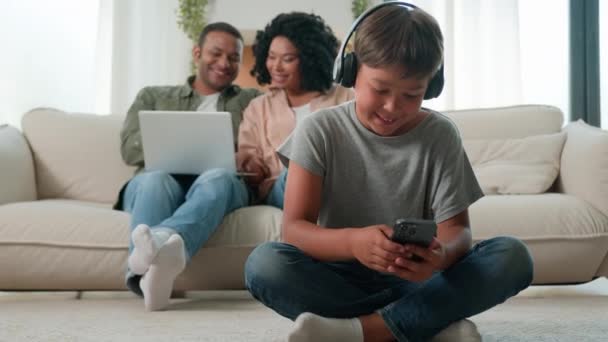 小男孩的孩子们戴着耳机在手机上玩电子游戏快乐的多种族妈妈和学龄前的孩子们在家里用各种各样的设备 家长们在网上购物 沙发上还带着笔记本电脑 — 图库视频影像