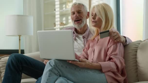 幸せな高齢者既婚カップル男性女性の顧客は インターネットの電子バンキングサービスでクレジットカードの安全な即時支払いと一緒に自宅で話すラップトップを使用してオンラインで購入を作成する抱擁 — ストック動画
