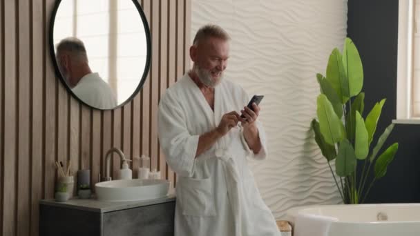 コーカサスハッピー 幸せな老人は浴室の年配の年配の男性で浴室の朝の衛生を見て携帯電話の読書良いニュース スマートフォンの驚いた驚きの携帯電話の達成 — ストック動画