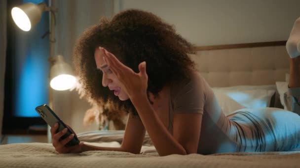 アフリカ系アメリカ人の若い民族の女性が心配する不安な少女は 夜の寝室でベッドに横たわっている携帯電話で悪いニュースを読んで悲しみ スマートフォンエラー通知を使用して女性に衝撃を与え ゲームを失う — ストック動画